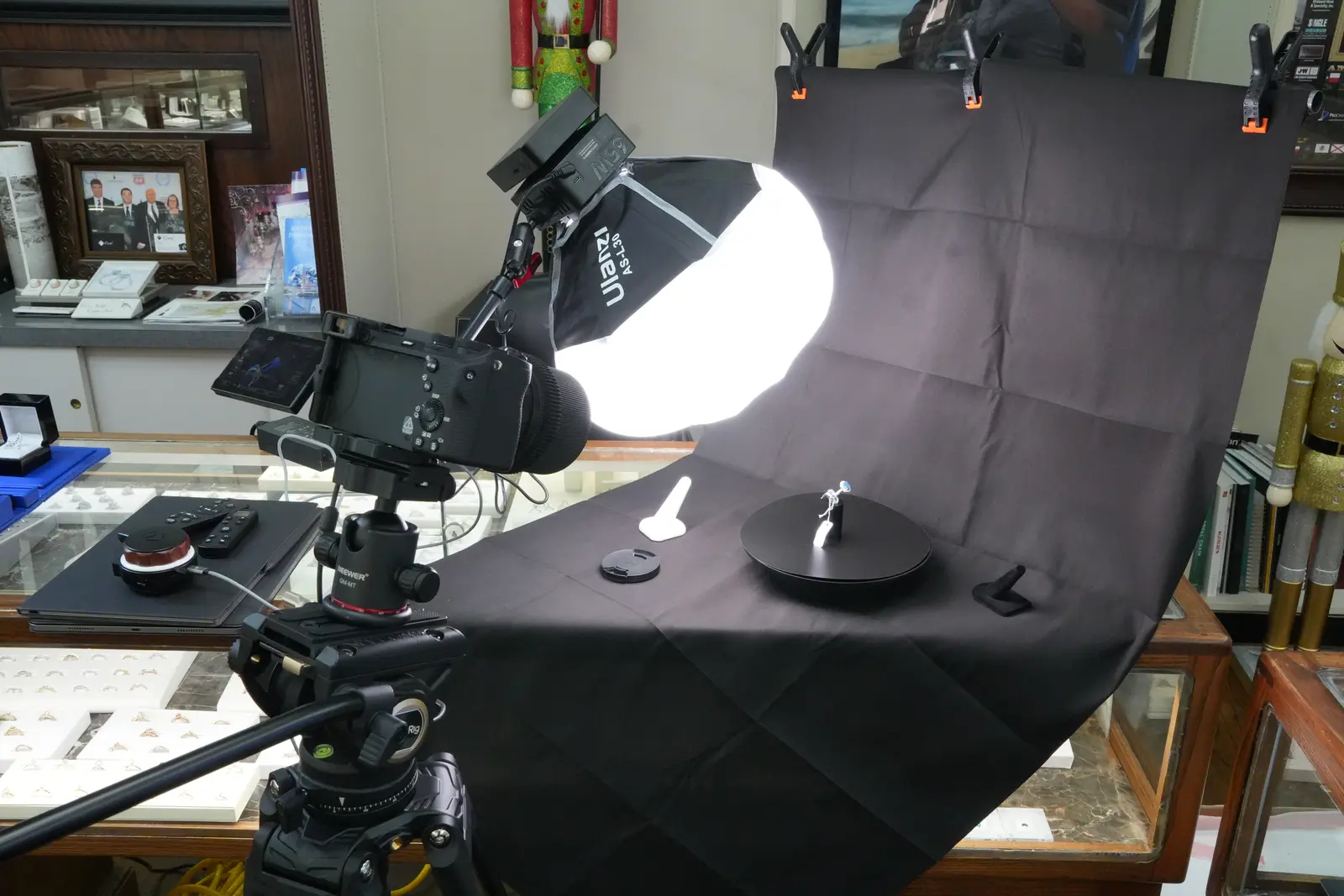 A ring on a black turntable in front of a black backdrop, the camera and softbox set up in a jewelry store with display cases behind