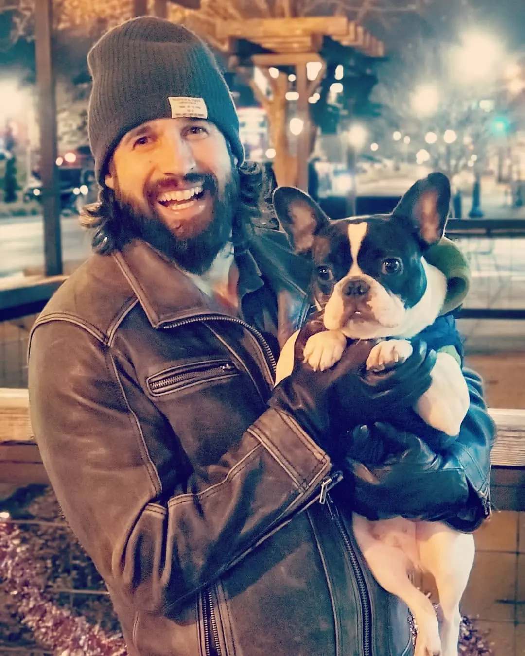 Mitch in a beanie and leather jacket holding his black and white French bulldog, with string lights behind them