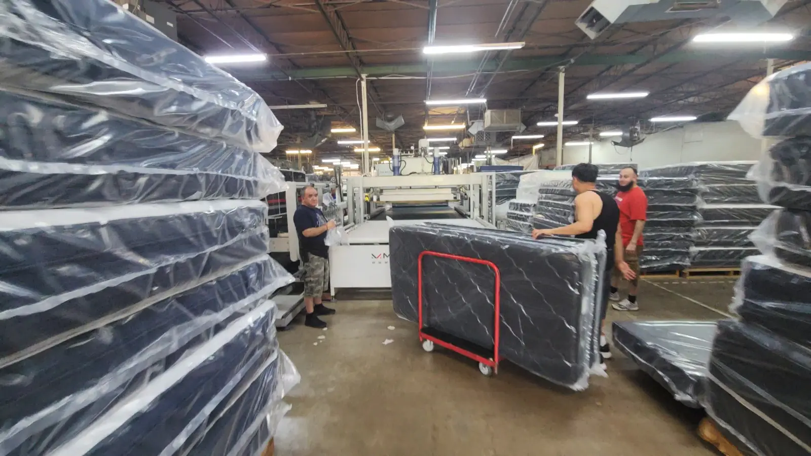 Workers moving plastic-wrapped mattresses past tall stacks on the Golden Mattress factory floor in Dallas